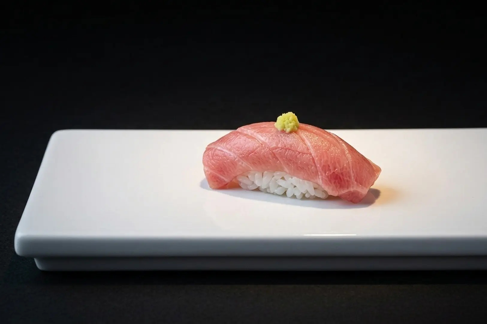 Single piece of tuna nigiri sushi with wasabi on white rectangular plate.