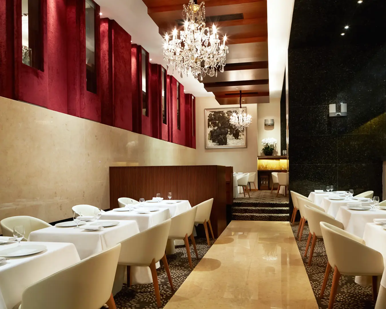 Elegant restaurant interior with white tablecloths and modern chairs. Chandeliers hang from a wood-paneled ceiling. Red velvet wall accents add warmth.