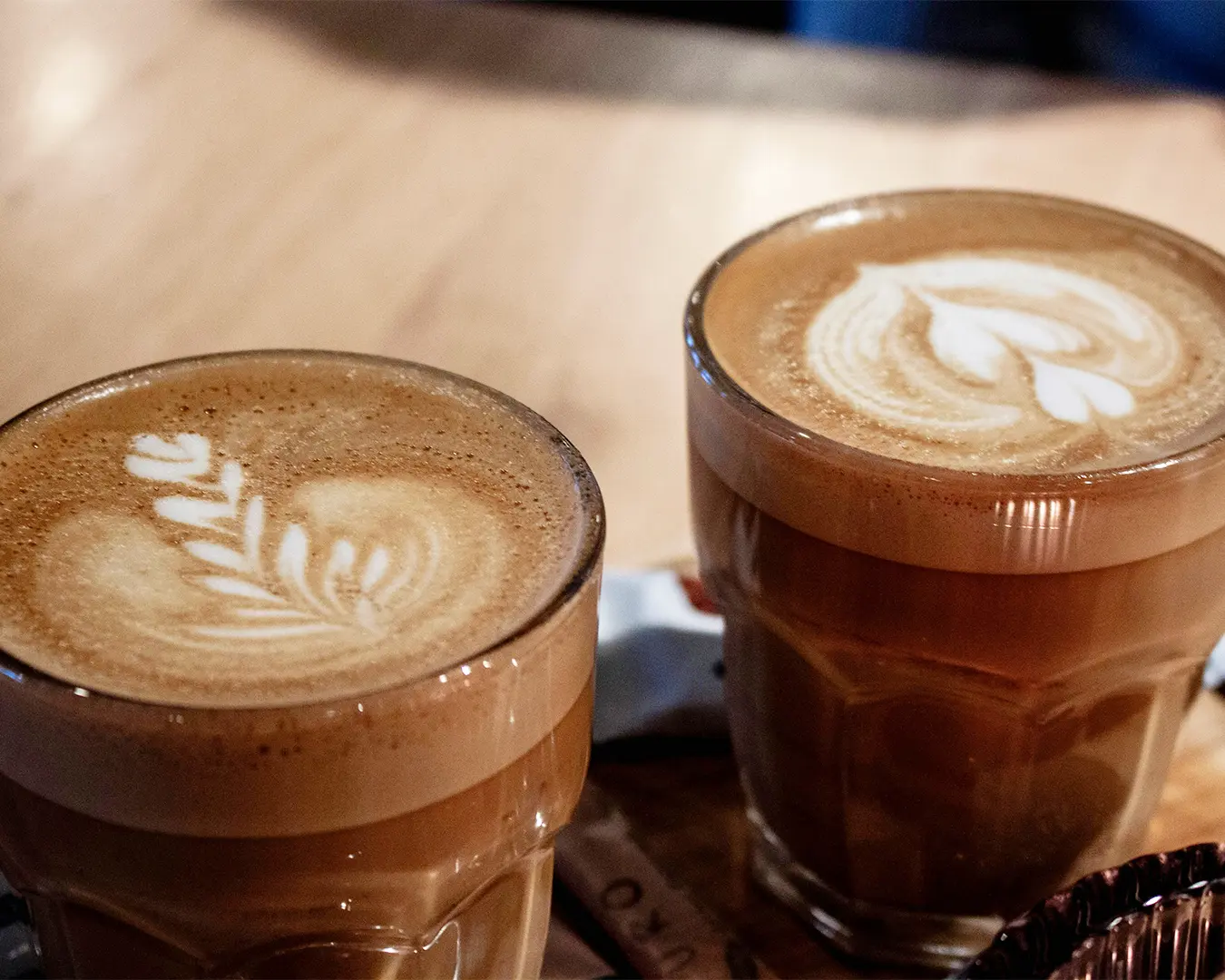 Two glass cups filled with lattes on a wooden table, featuring intricate latte art designs. The scene conveys a cozy and warm coffee shop ambiance.
