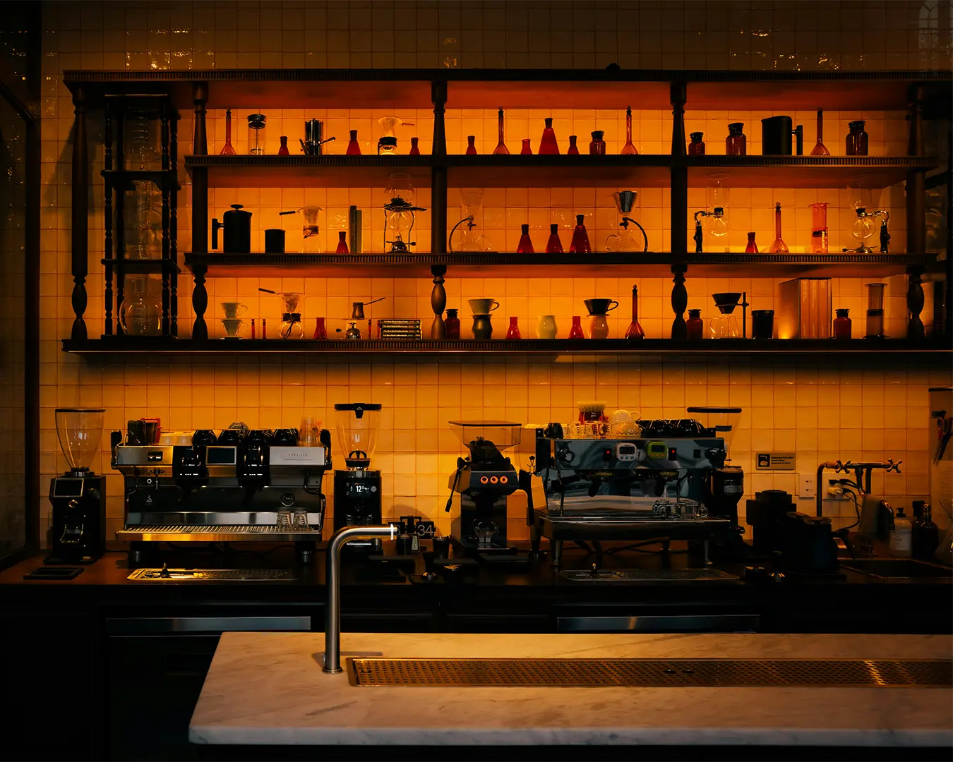 Cozy cafe scene with a dimly lit coffee bar, featuring espresso machines and a warm glow from an array of glassware on backlit shelves.