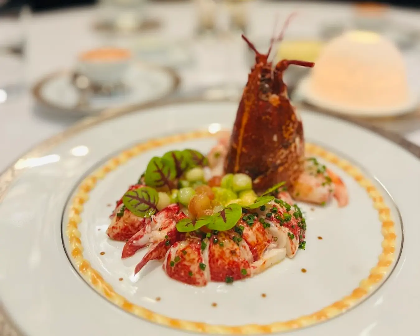 Elegant lobster dish on a white plate, garnished with greens and colorful sauce. The presentation is artistic, conveying luxury and sophistication.