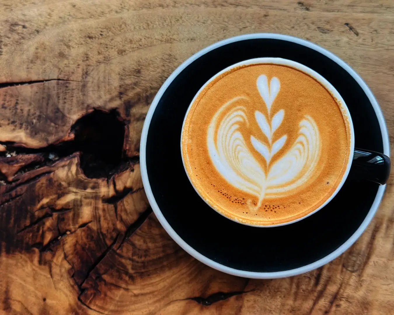 A latte with leaf art in a black cup on a rustic wooden table. The smooth frothy latte contrasts with the textured wood, creating a warm, inviting tone.