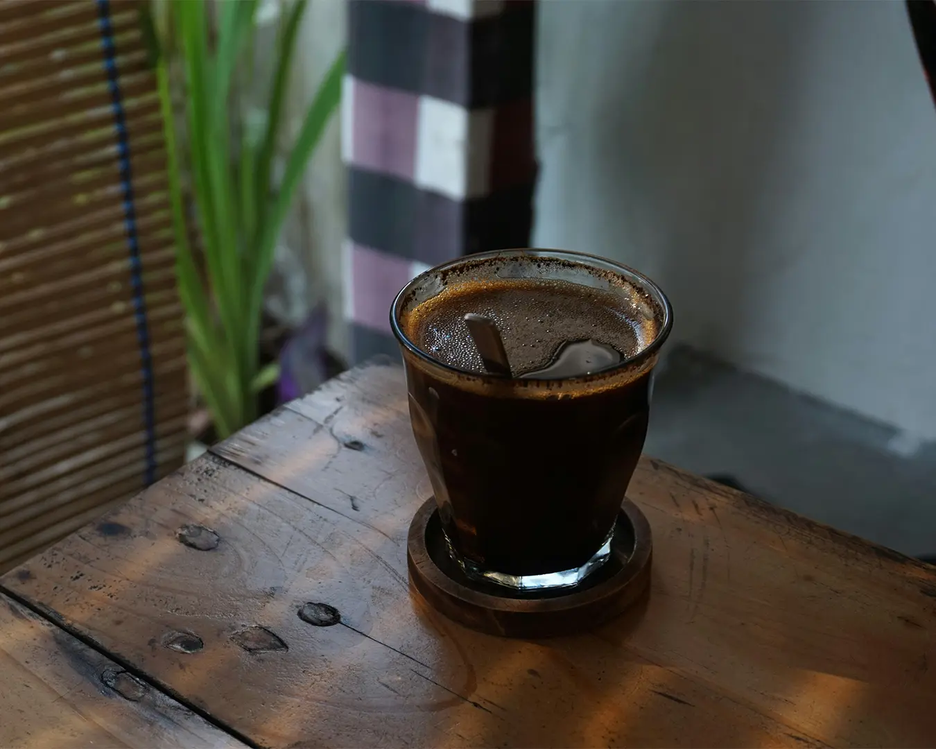 A glass of black coffee sits on a rustic wooden table, with a bamboo blind and leafy green plant in the background, creating a cozy, relaxed atmosphere.
