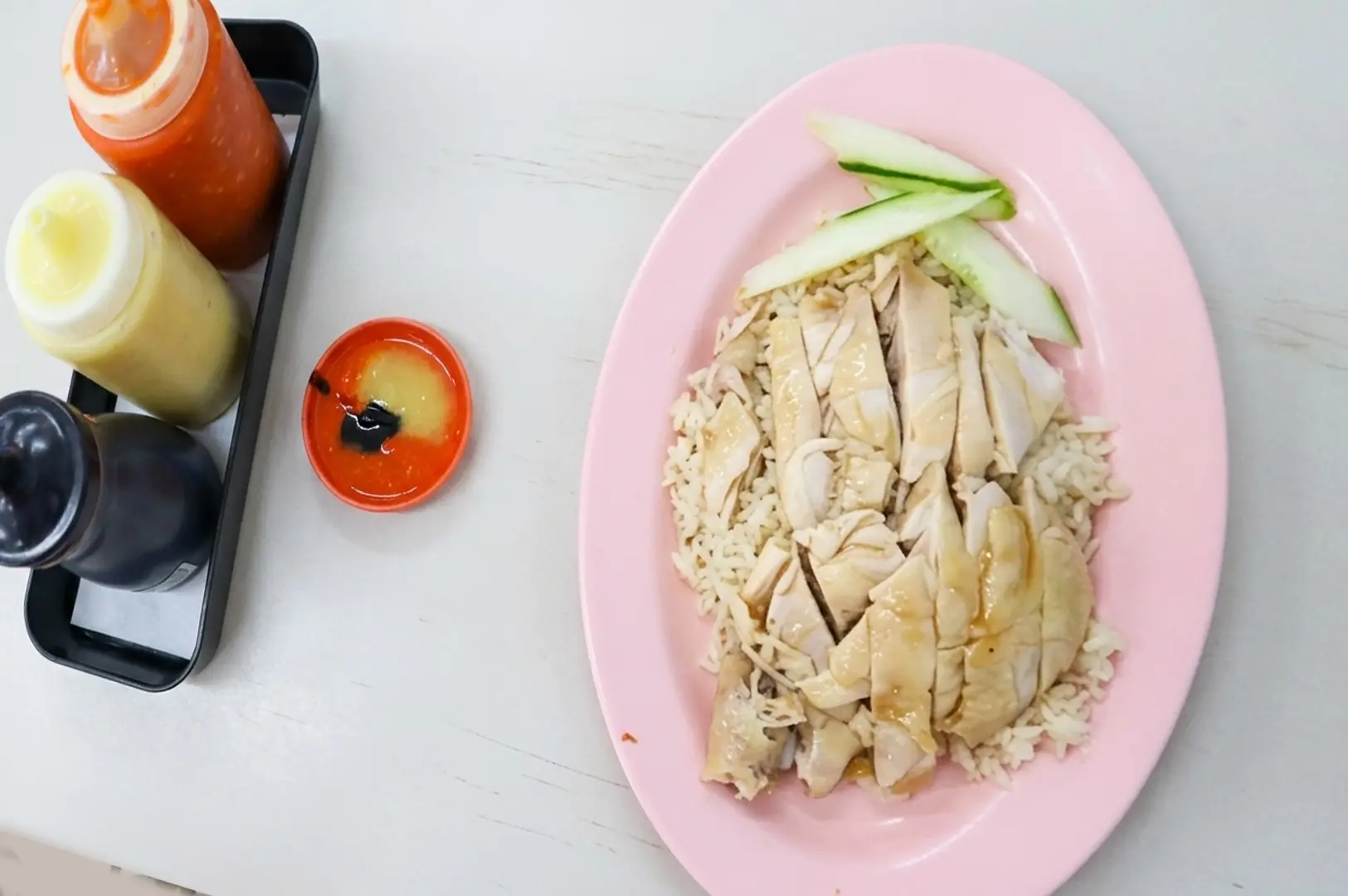 Hainanese chicken rice on pink plate with sliced cucumber and three condiment bottles