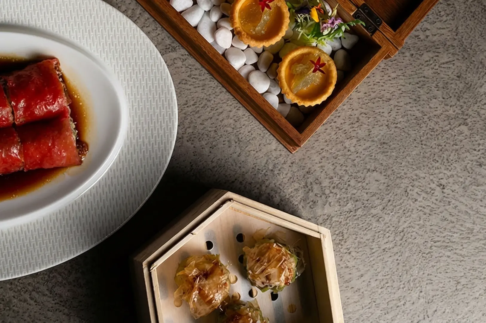 Elegant Chinese fine dining presentation with glazed char siu pork on textured white plate and artistic dim sum arrangements in wooden boxes with decorative white stones