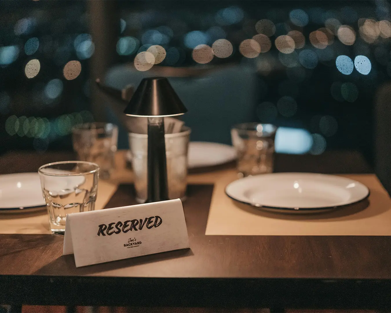 A dimly lit restaurant table set with plates and glasses, featuring a small lamp. A "Reserved" card is prominently displayed, with city lights blurred in the background.