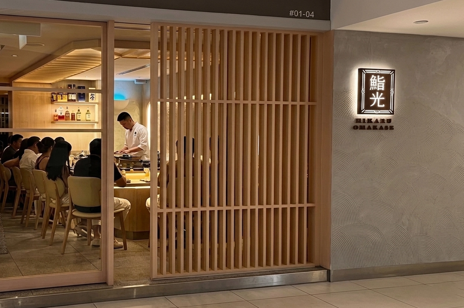 Hikaru Omakase restaurant entrance with wooden slat partition and chef preparing sushi at counter.