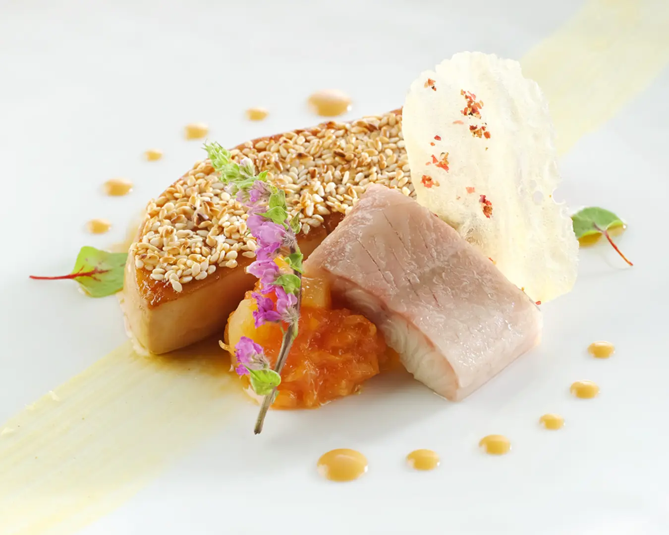 Gourmet dish on a white plate features sesame-crusted fish, a purple flower garnish, a slice of translucent crispy cracker, and vibrant orange sauce. Elegant presentation.