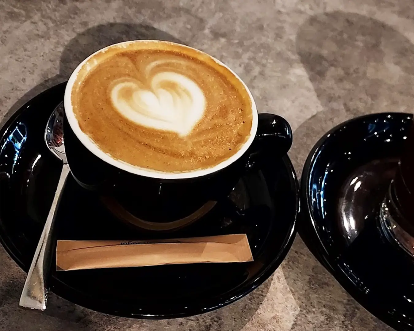 A cappuccino in a black cup on a saucer, featuring heart-shaped latte art. Next to it, a spoon and a sugar packet on a textured table surface.