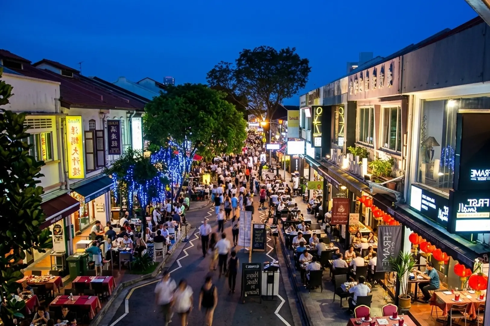 Busy Holland Village street at dusk with crowded outdoor dining, restaurants with patrons, and decorative blue lights on trees