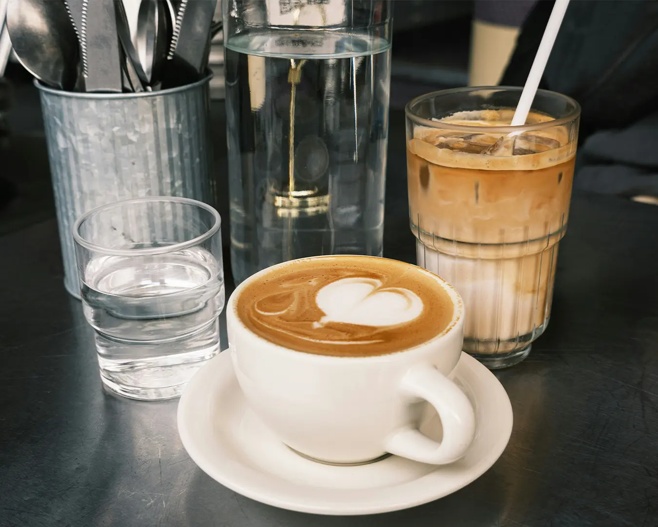 A cozy café scene with a white cup of cappuccino topped with heart-shaped latte art, a glass of iced coffee, a water glass, and cutlery in a tin holder.