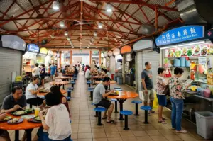 Busy hawker center with orange tables, blue stools, and diners eating at food stalls