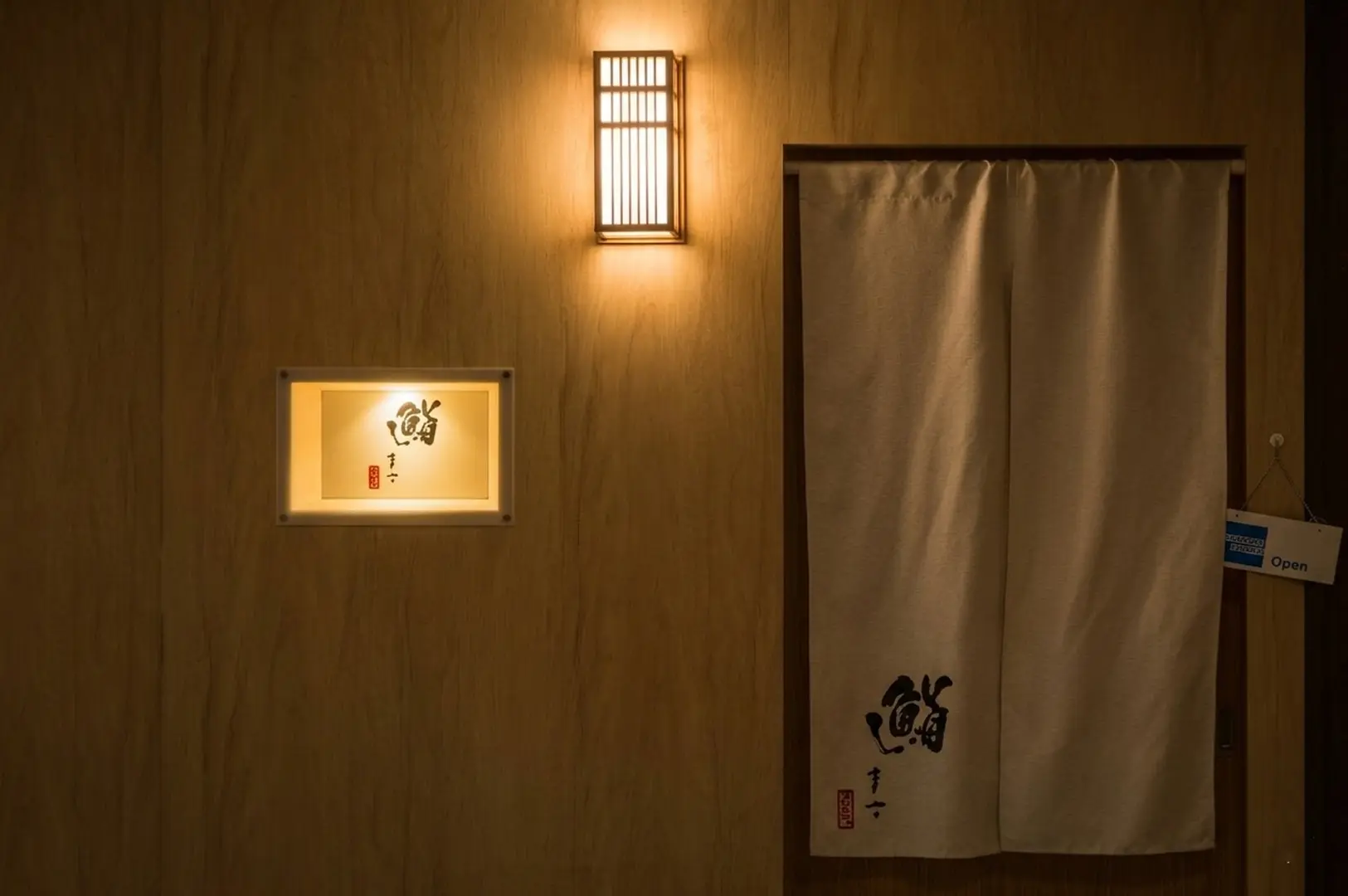 Entrance to the restaurant showing a white noren curtain with the calligraphy logo and an Open sign