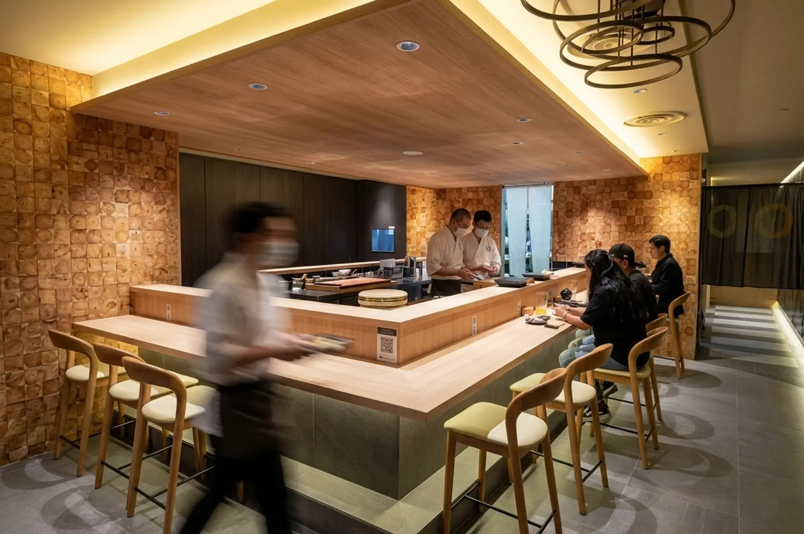Modern fine dining restaurant with wooden sushi counter, chefs in white uniforms preparing dishes, contemporary lighting, and elegant wood-paneled interio