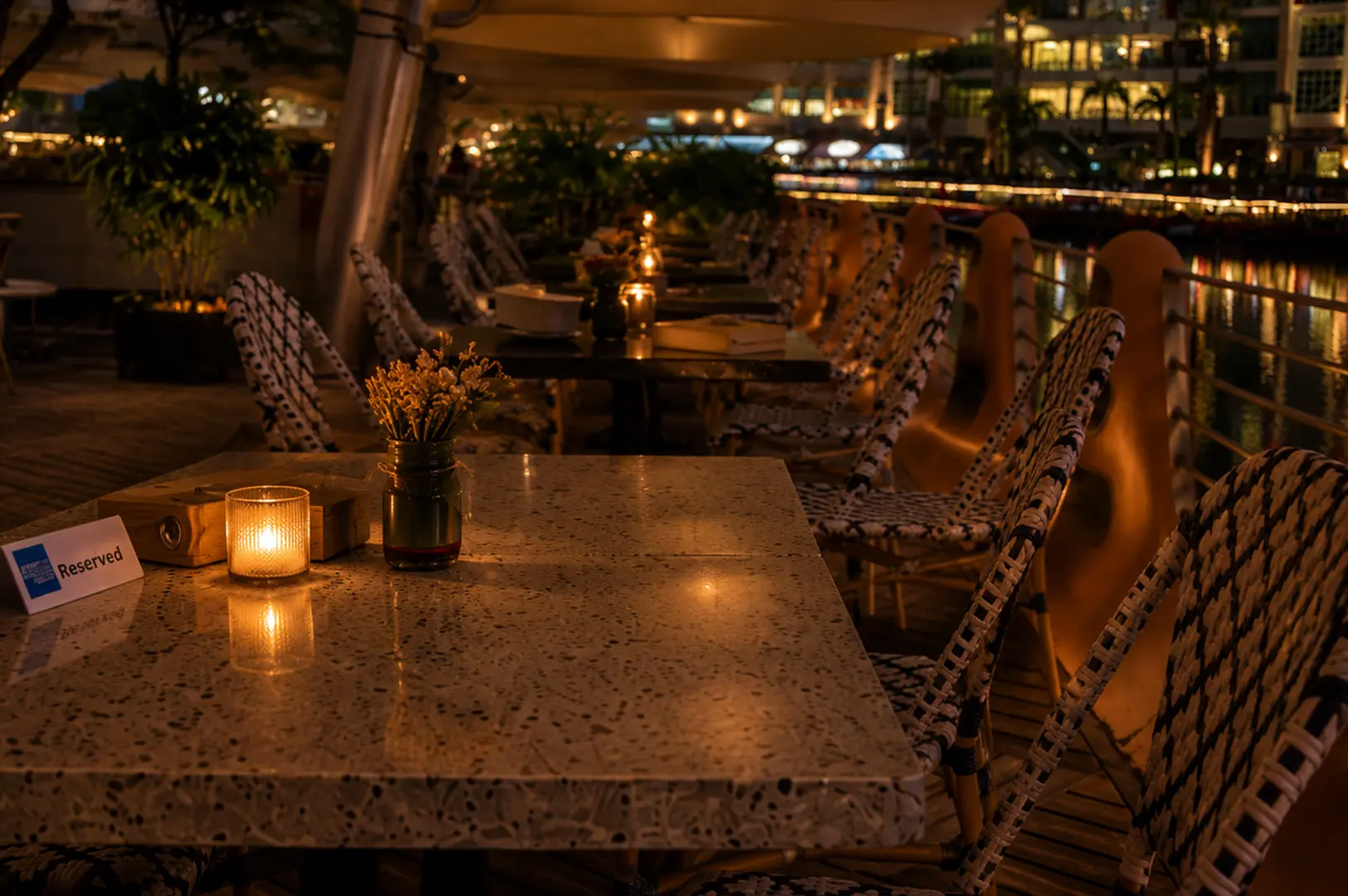 Romantic outdoor waterfront restaurant terrace with candlelit tables and city lights at night.