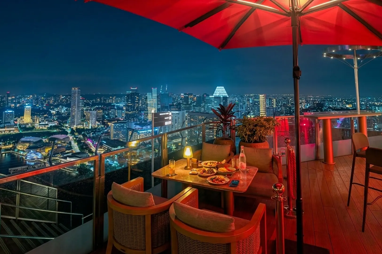 Rooftop dining table under red umbrella with panoramic night view of illuminated Singapore skyline