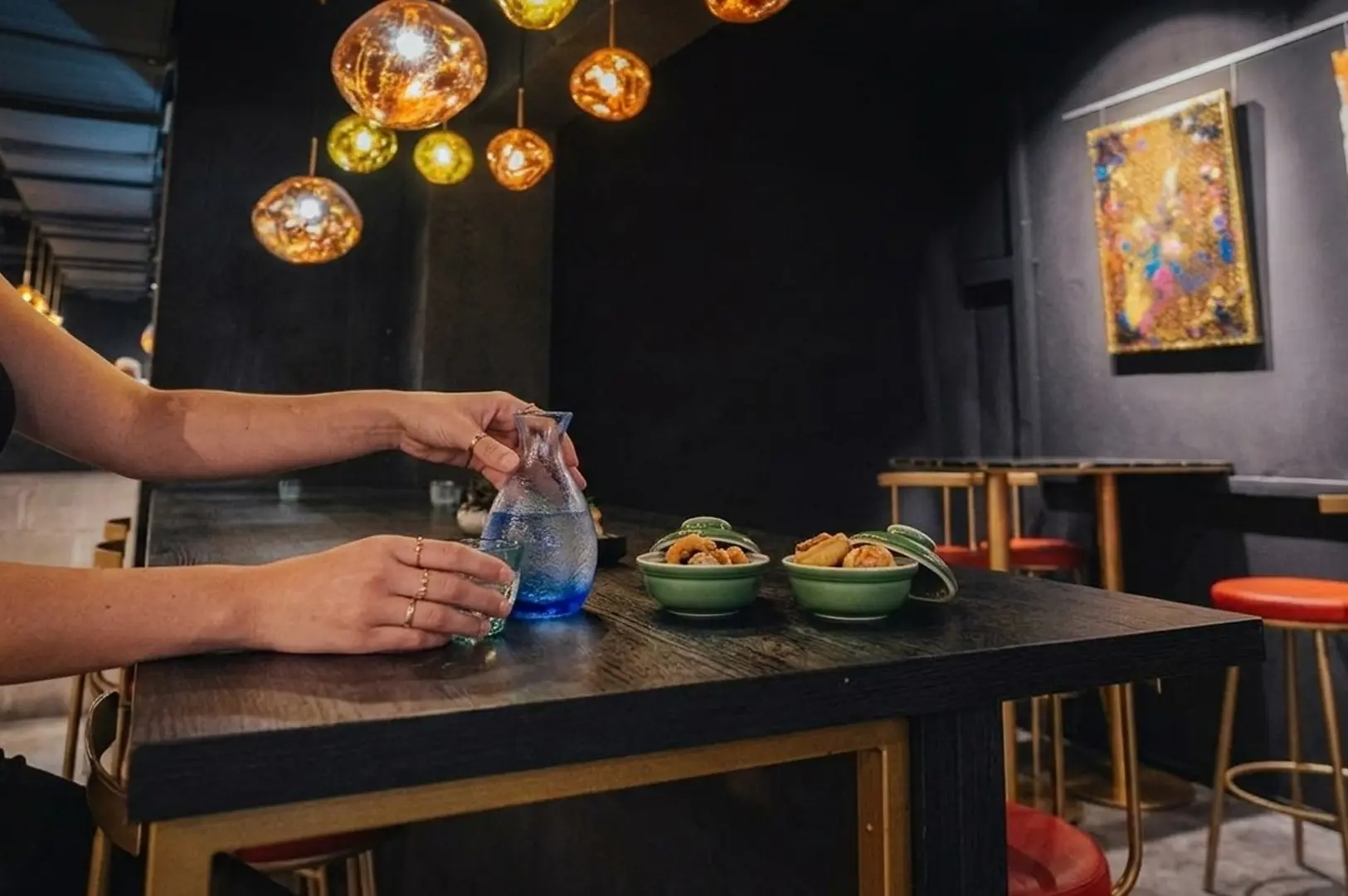Person pouring blue cocktail at intimate Telok Ayer wine bar with small bites in green ceramic bowls under warm pendant lights