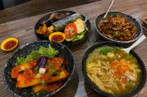 Four Indonesian dishes including grilled chicken, sambal eggplant, tempeh, and chicken noodle soup served in speckled ceramic bowls on a wooden table
