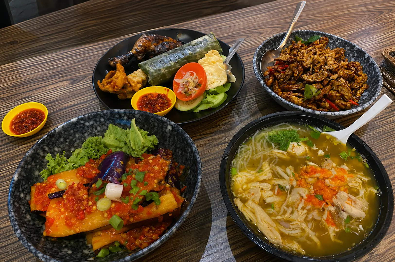 Four Indonesian dishes including grilled chicken, sambal eggplant, tempeh, and chicken noodle soup served in speckled ceramic bowls on a wooden table