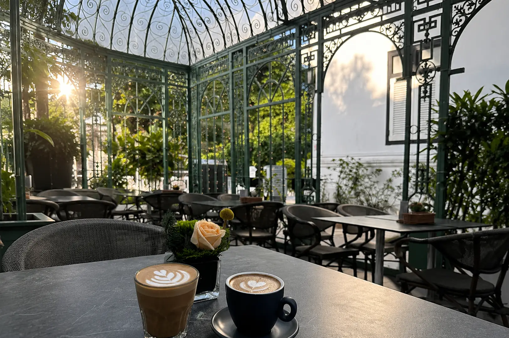 Two lattes with heart art on a black table at an outdoor cafe with green metal gates and plants