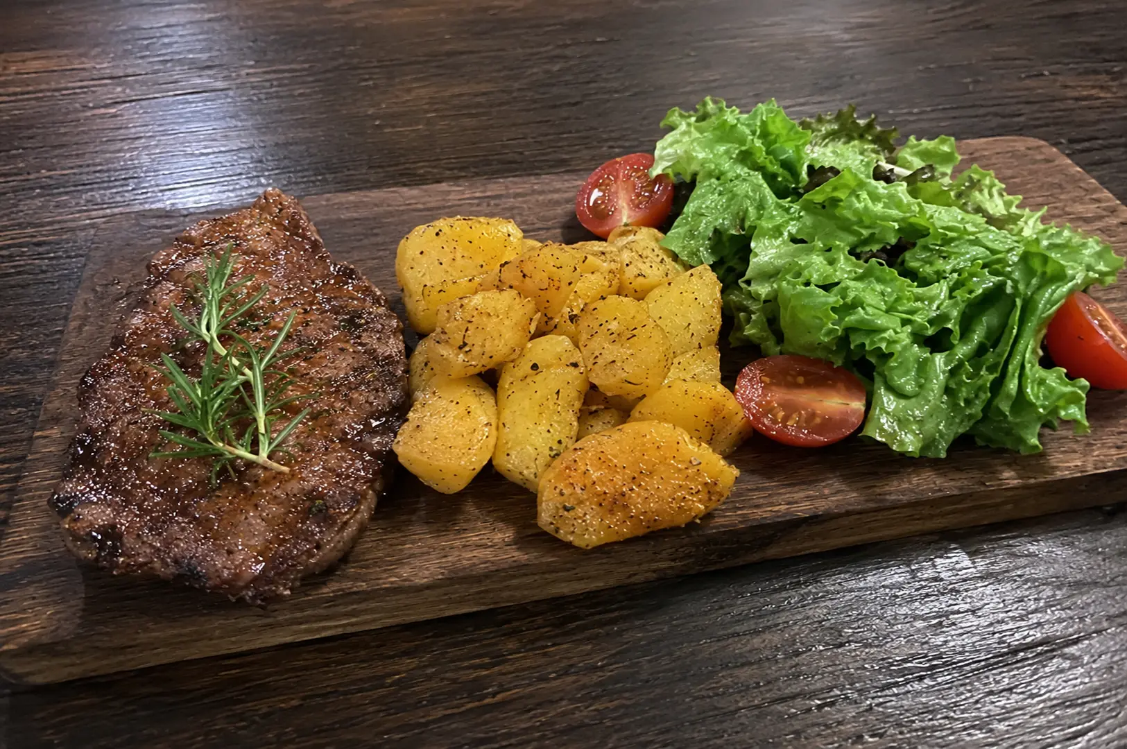 Grilled steak garnished with rosemary sprig served with herb-roasted potatoes and fresh salad on a rustic wooden serving board