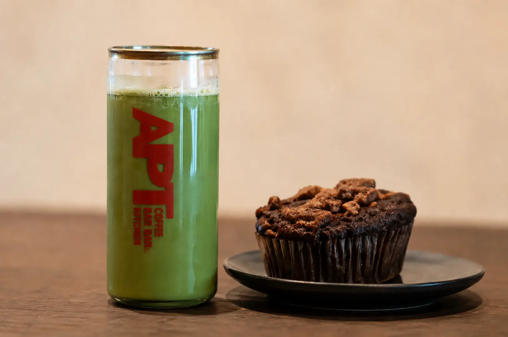 A tall glass of matcha drink with APT Coffee Bar & Kitchen branding next to a chocolate walnut muffin on a black plate