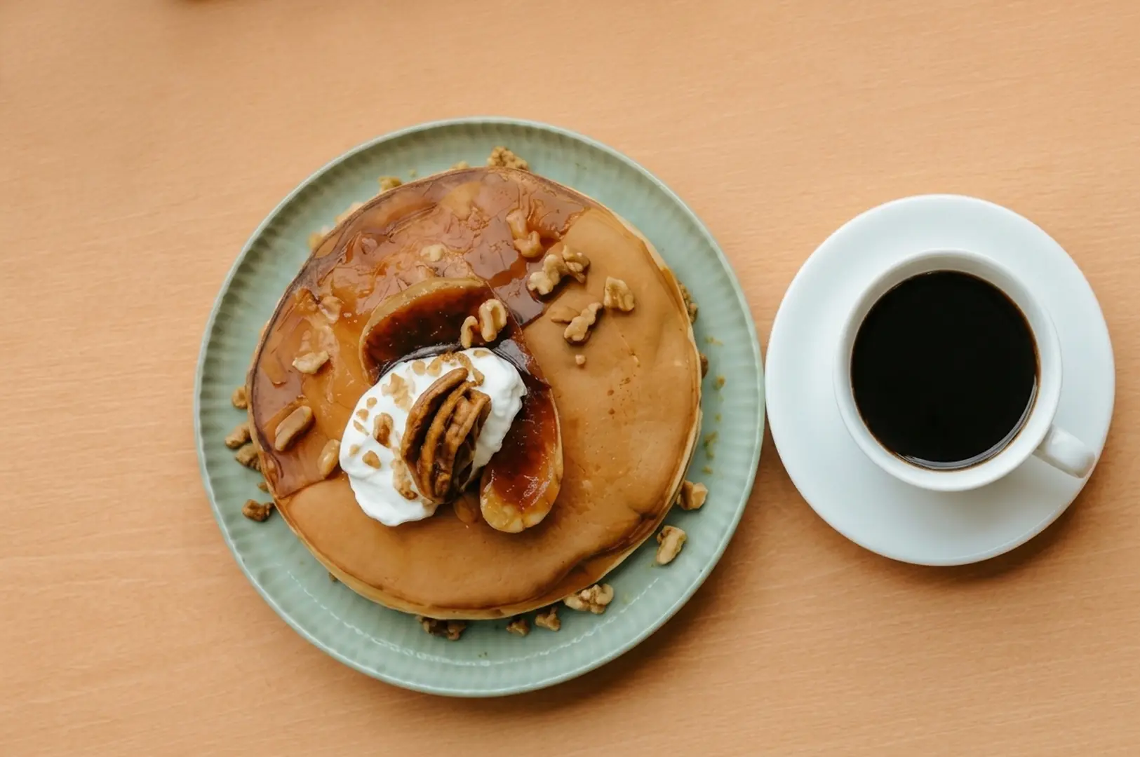 Stack of pancakes with caramelized bananas, walnuts, and cream on a green plate next to a cup of black coffee