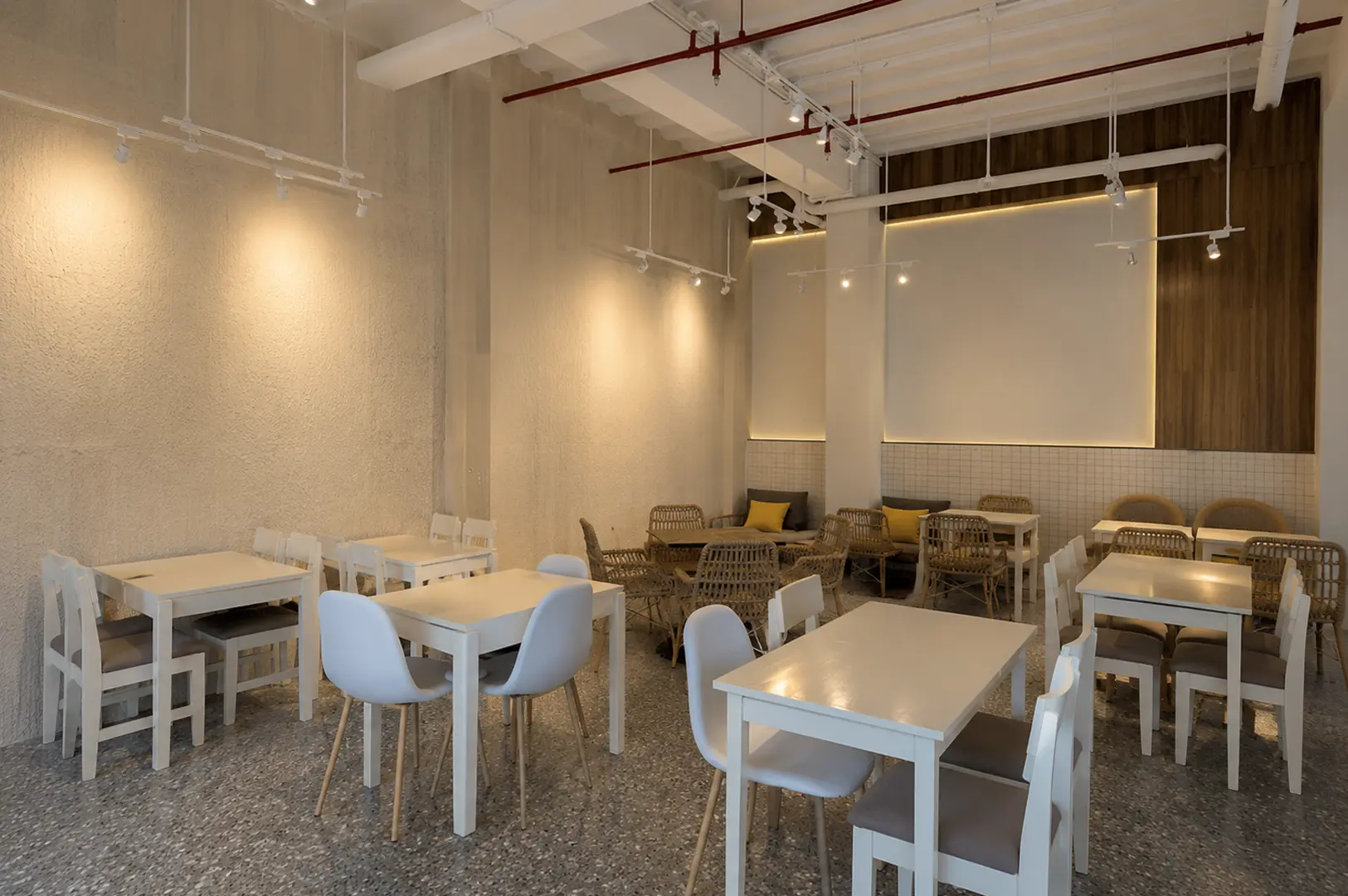 Spacious cafe dining area with white tables, rattan seating, exposed ceiling pipes, and a large projection screen