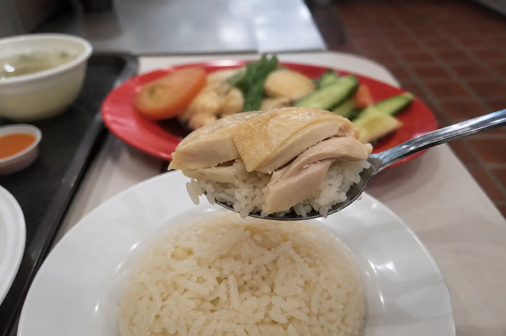 Close-up of a spoonful of Hainanese chicken rice with a full plate and soup in the background