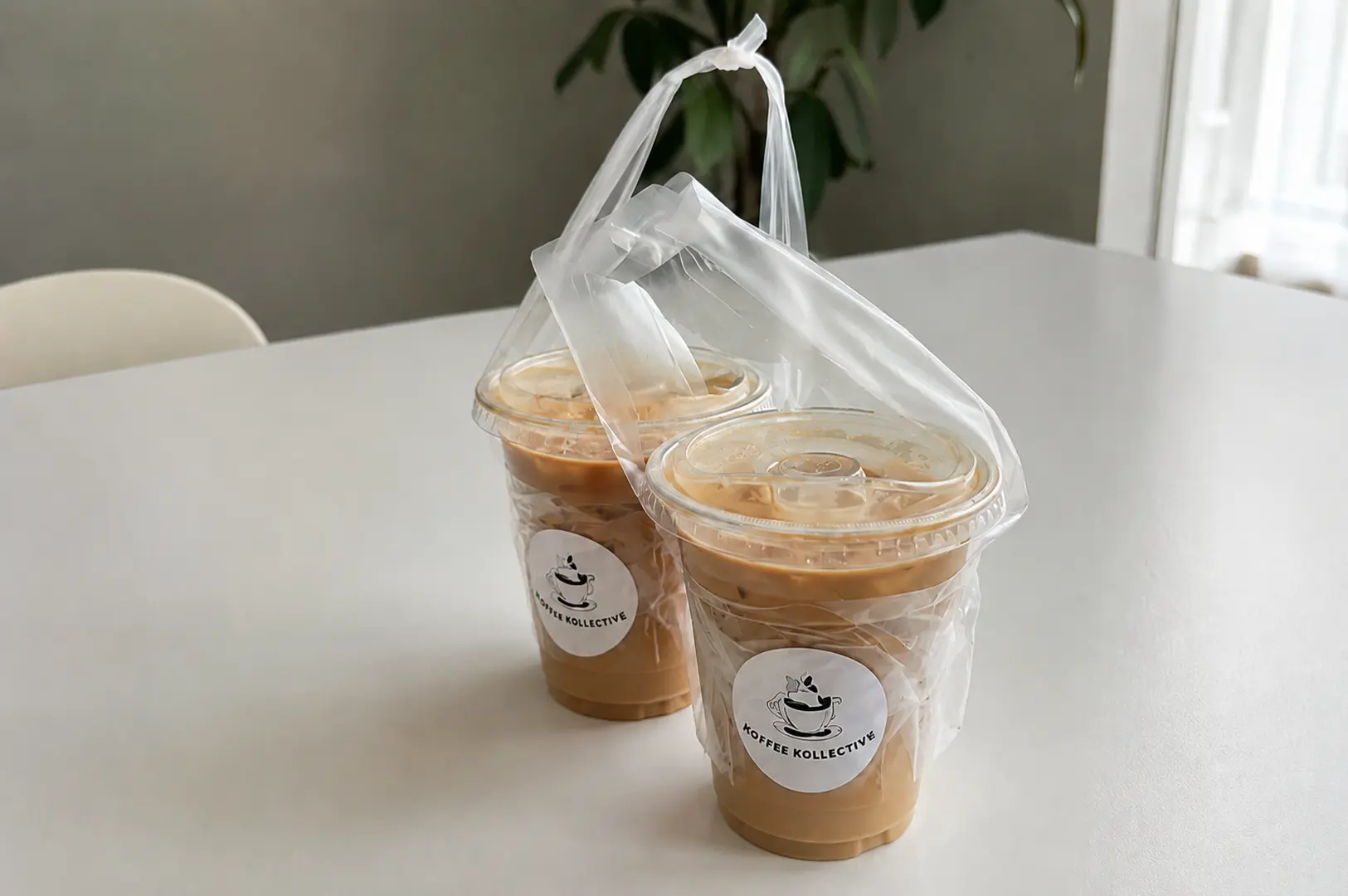 Two iced coffee drinks in plastic cups with Koffee Kollective logo stickers carried in a transparent plastic bag on a white table