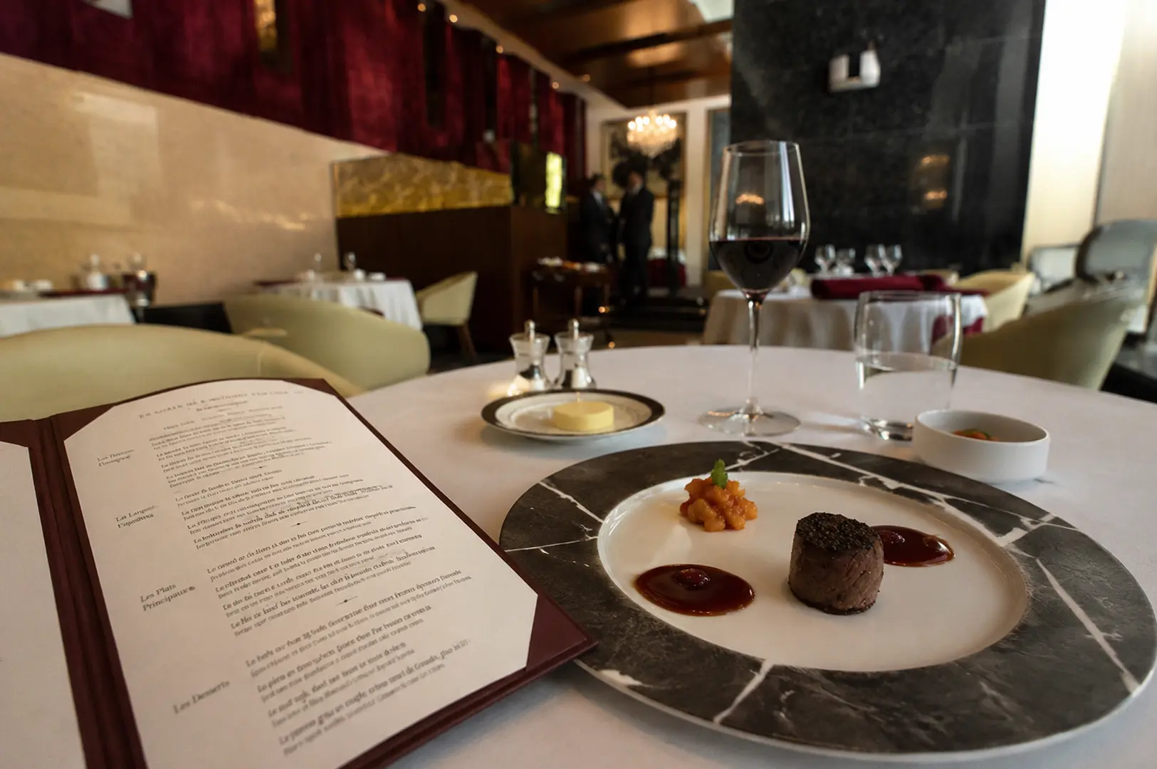 Fine dining table setting with a plated steak dish, red wine, and an open menu in an elegant restaurant with red velvet curtains.