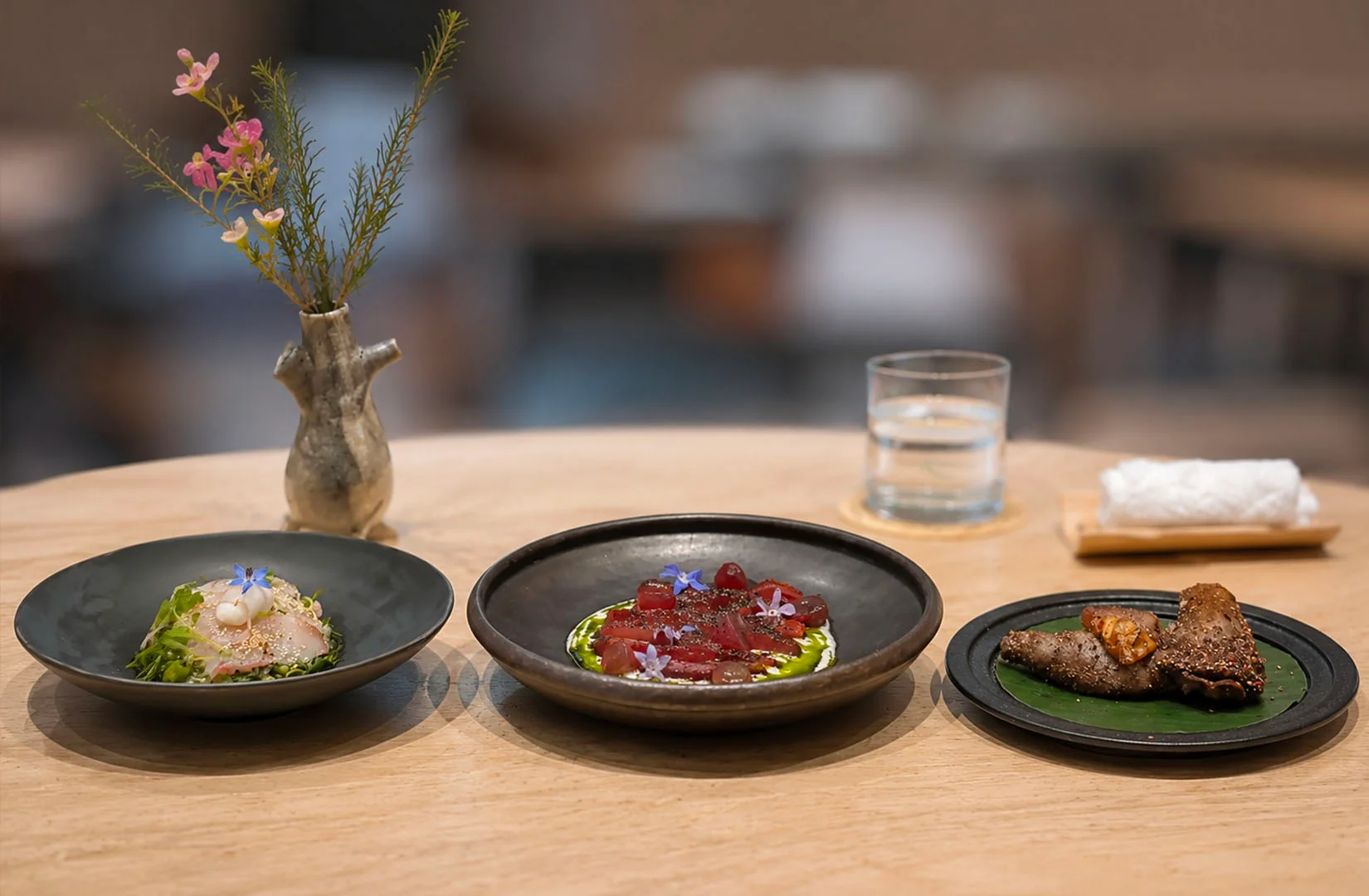 Three artfully plated fine dining dishes featuring sashimi, beet carpaccio with edible flowers, and grilled meat on a stone table