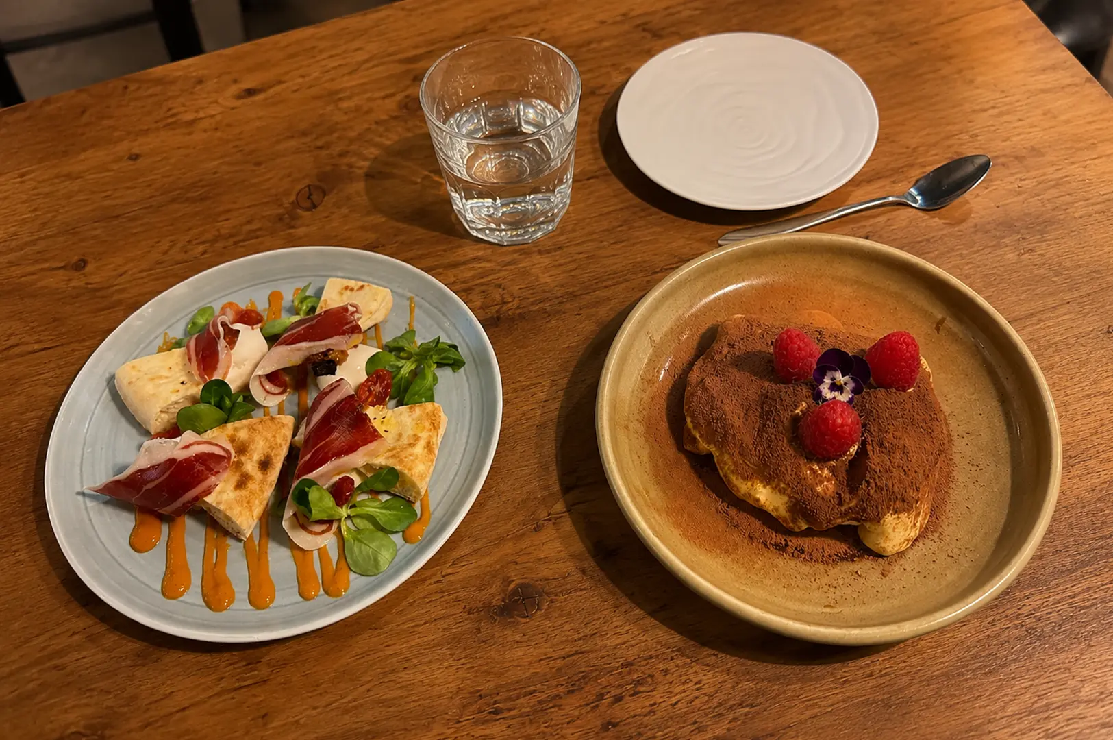 Italian appetizer platter with prosciutto-wrapped bread and orange sauce alongside tiramisu topped with raspberries and edible flowers on a wooden table