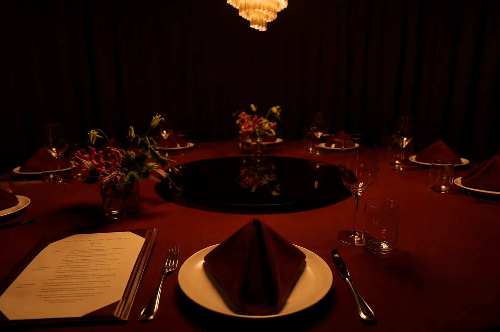 Dimly lit private dining room featuring a large round table with a maroon tablecloth, elegant place settings, and floral centerpieces