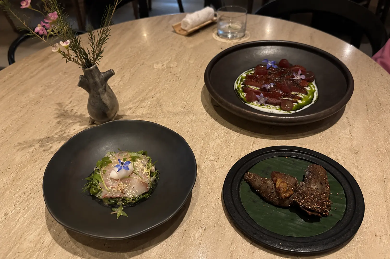 Three artfully plated fine dining dishes featuring sashimi, beet carpaccio with edible flowers, and grilled meat on a stone table