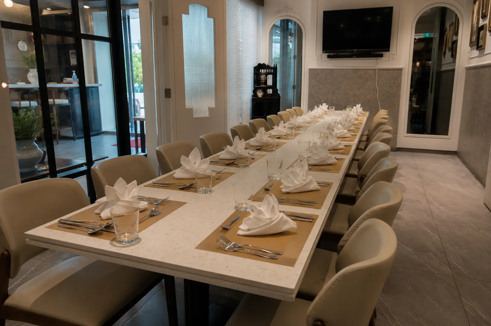Long rectangular private dining table set for a large group with white napkins, silverware, and beige chairs in a bright room