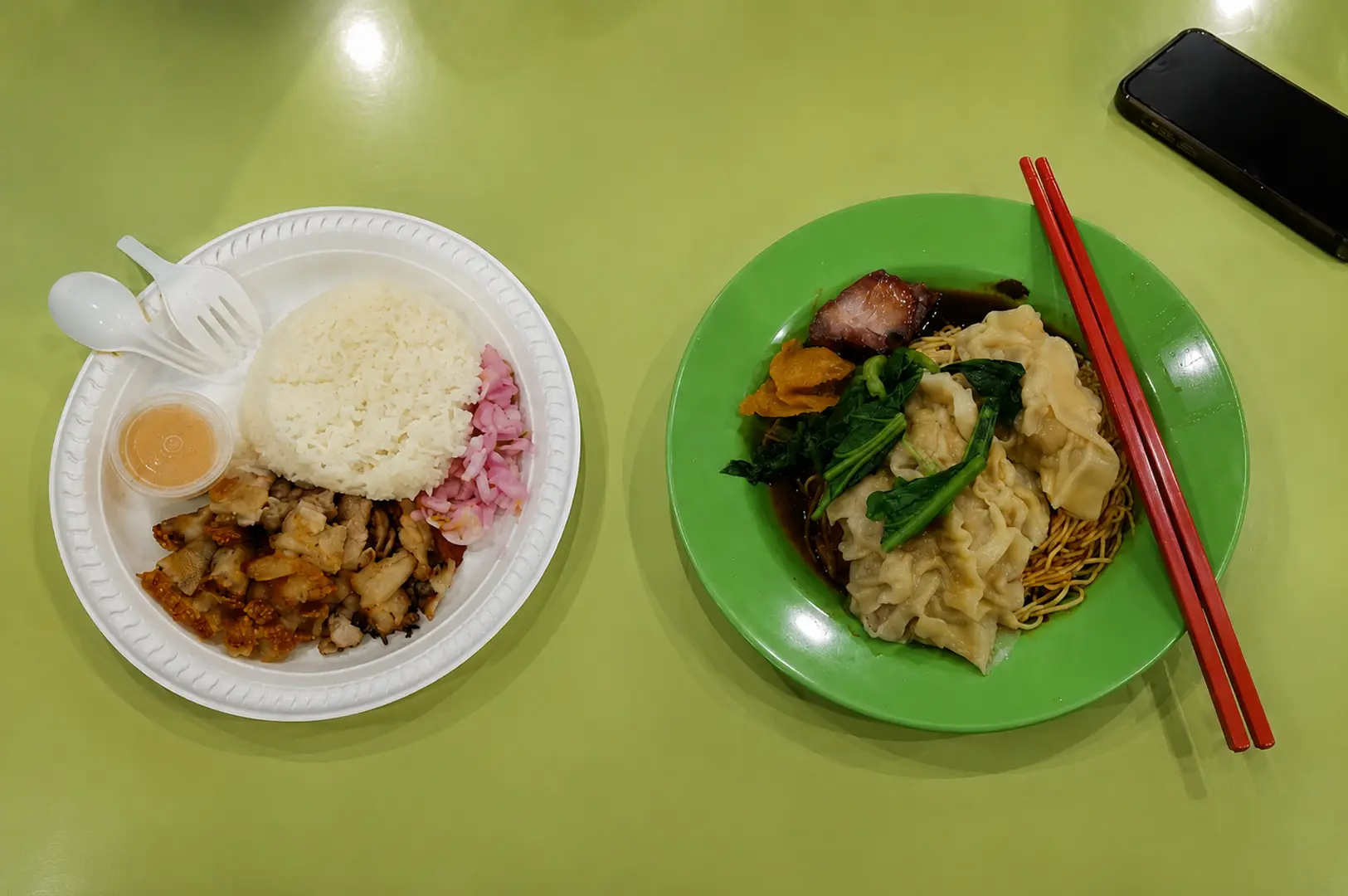 Two plates of hawker food featuring crispy pork rice and wonton noodles with char siu on a bright green table at a local food center