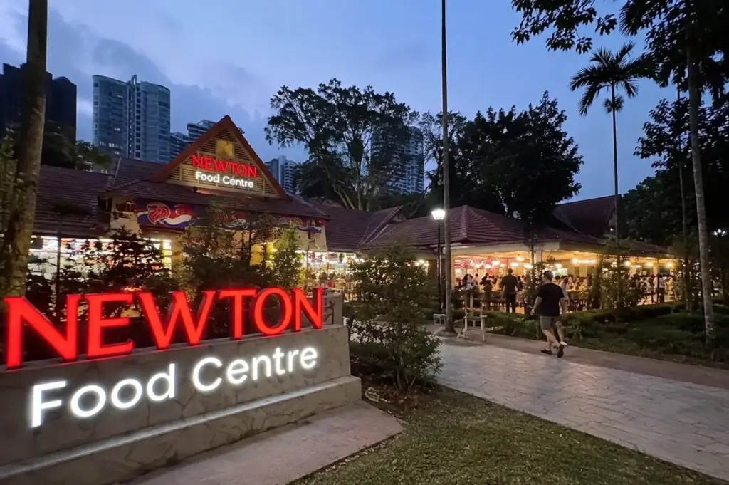 Evening view of Newton Food Centre with its glowing entrance sign and bustling open-air dining area.