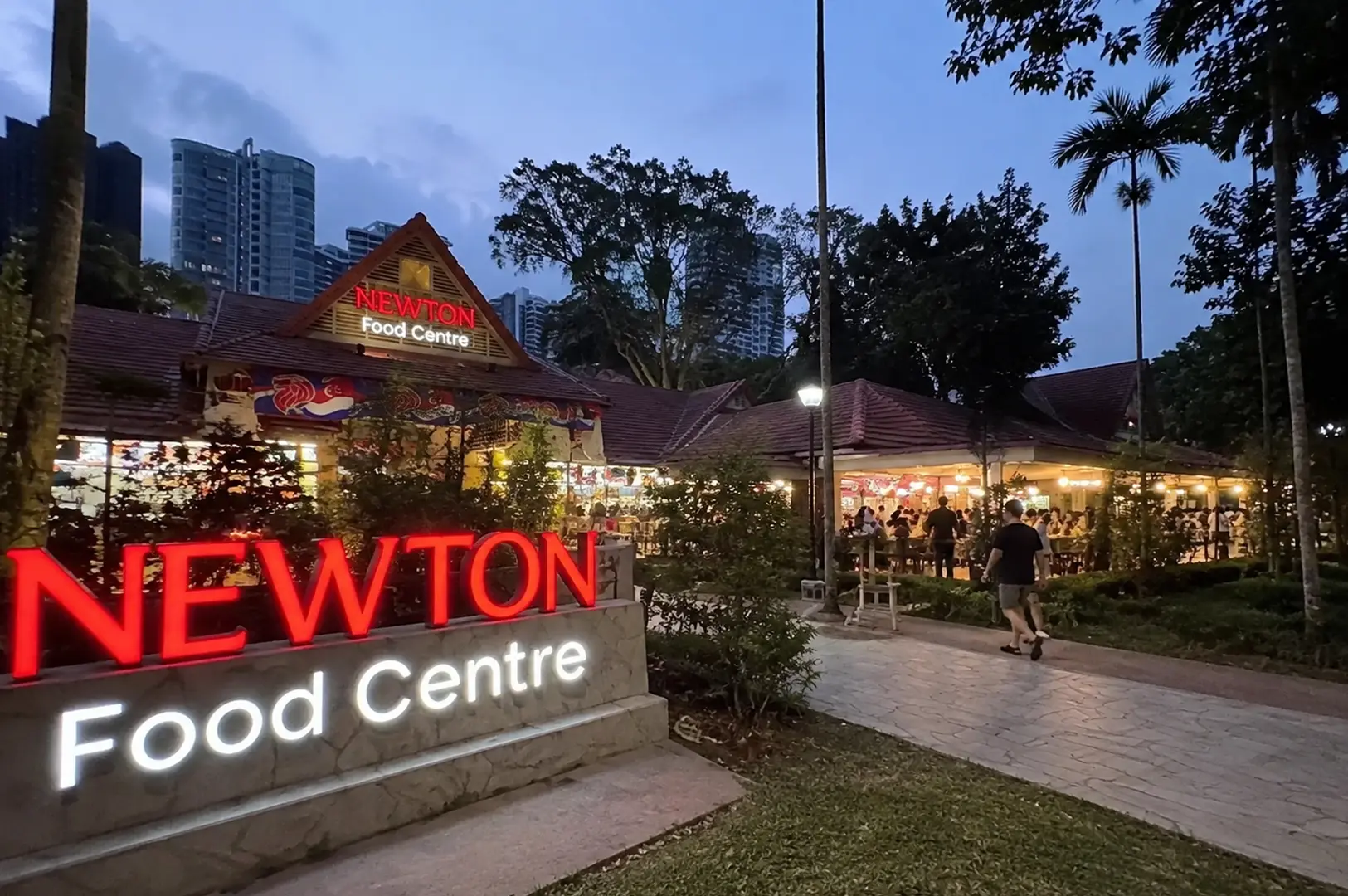 Evening view of Newton Food Centre with its glowing entrance sign and bustling open-air dining area.