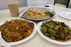 A hawker table spread featuring prawn noodles, spicy fried noodles, and stir-fried petai beans with meat, alongside an iced drink