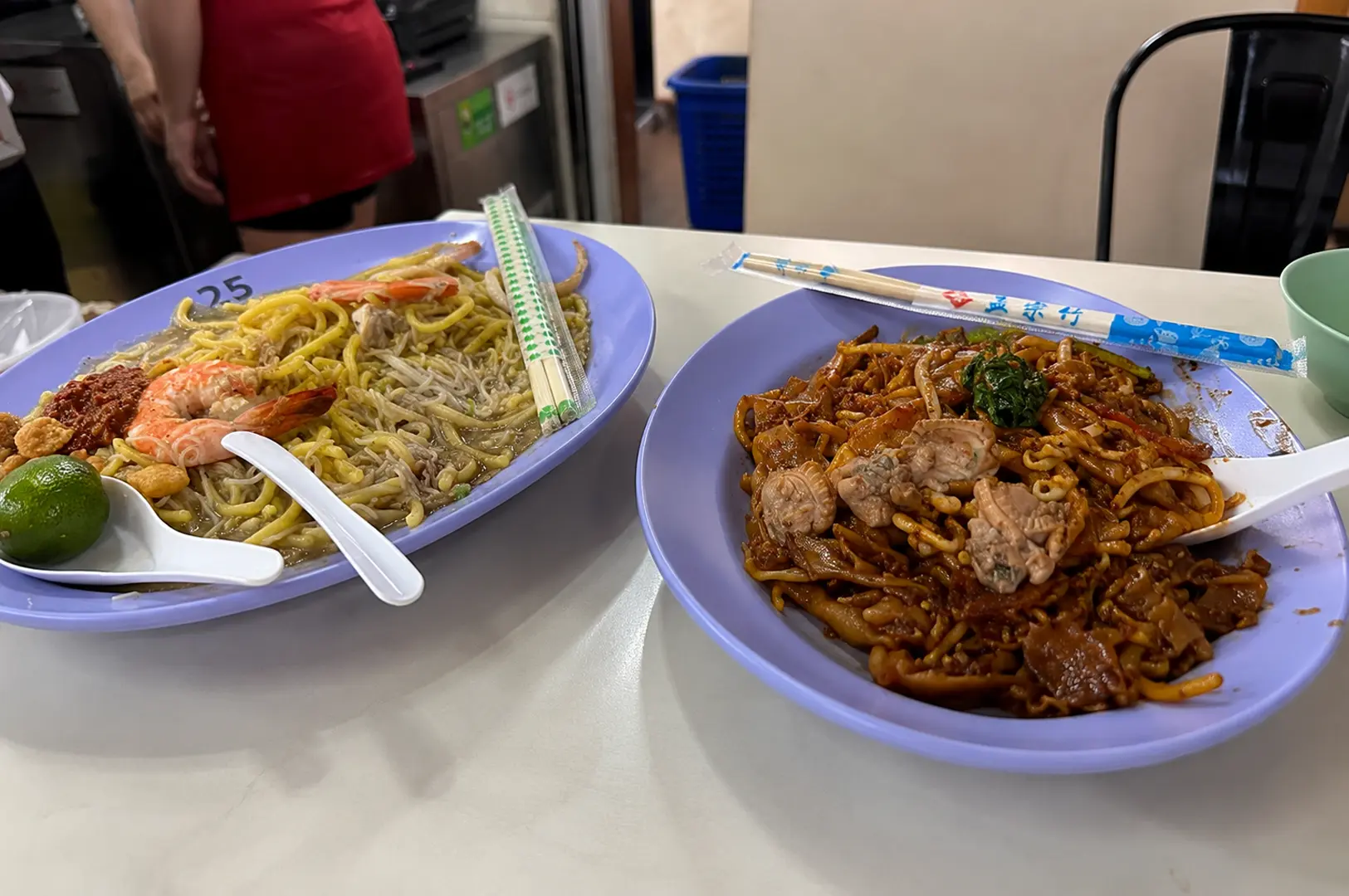 Two purple plates of hawker noodles - prawn mee with fresh prawns and yellow noodles alongside stir-fried flat noodles with meat and vegetables