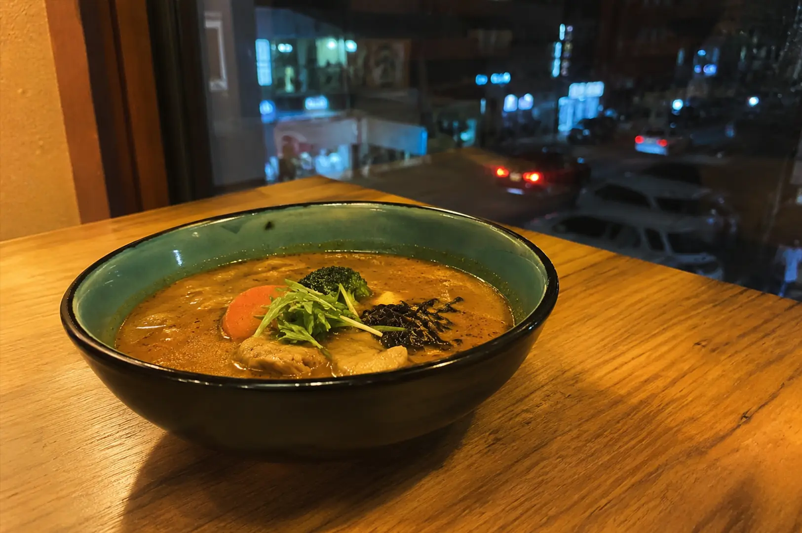 Bowl of Hokkaido-style soup curry served on a wooden table beside a window overlooking a quiet night street.