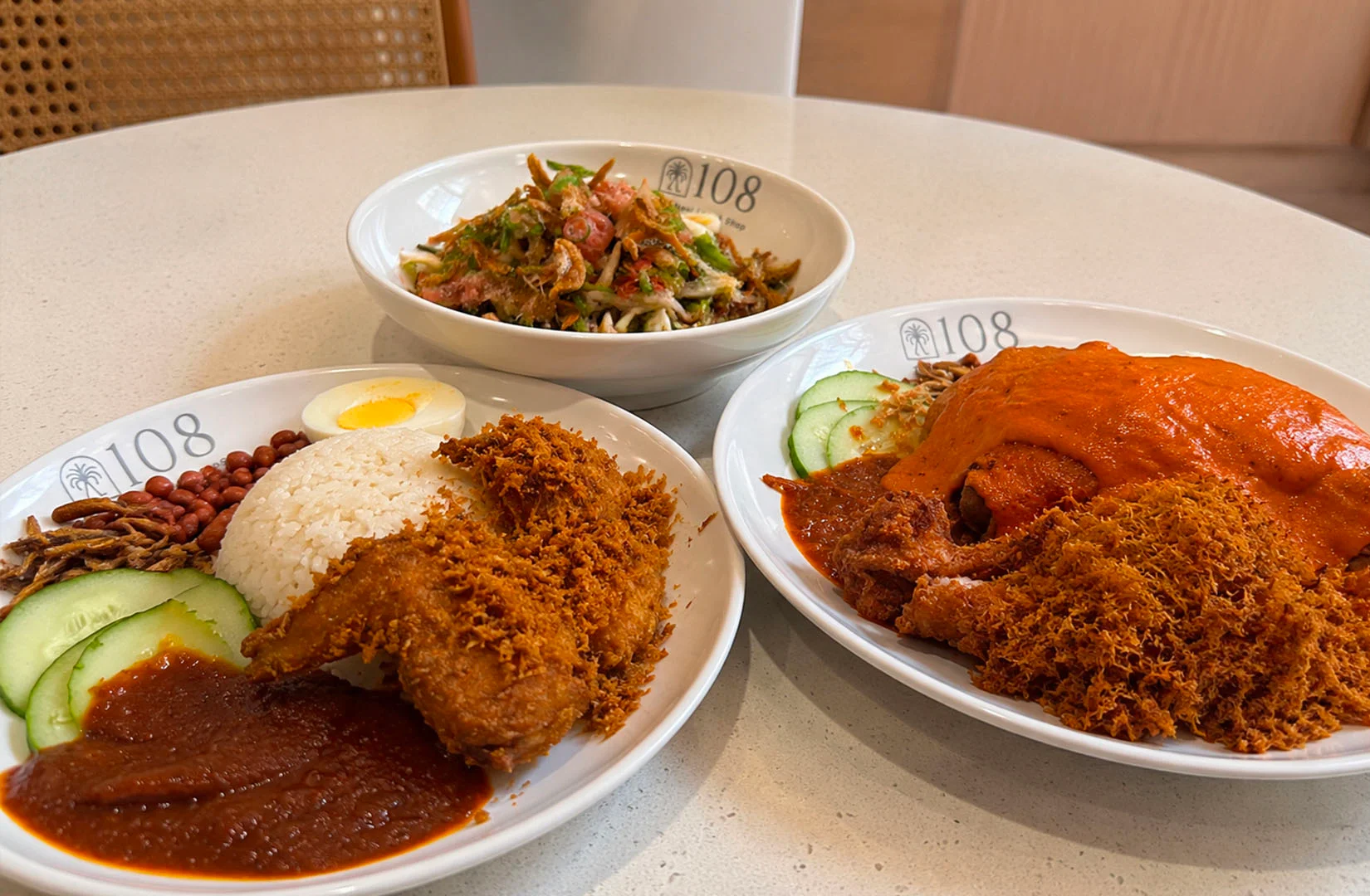 Plates of Singapore-style nasi lemak with fried chicken, rice, cucumber, peanuts, and sambal.