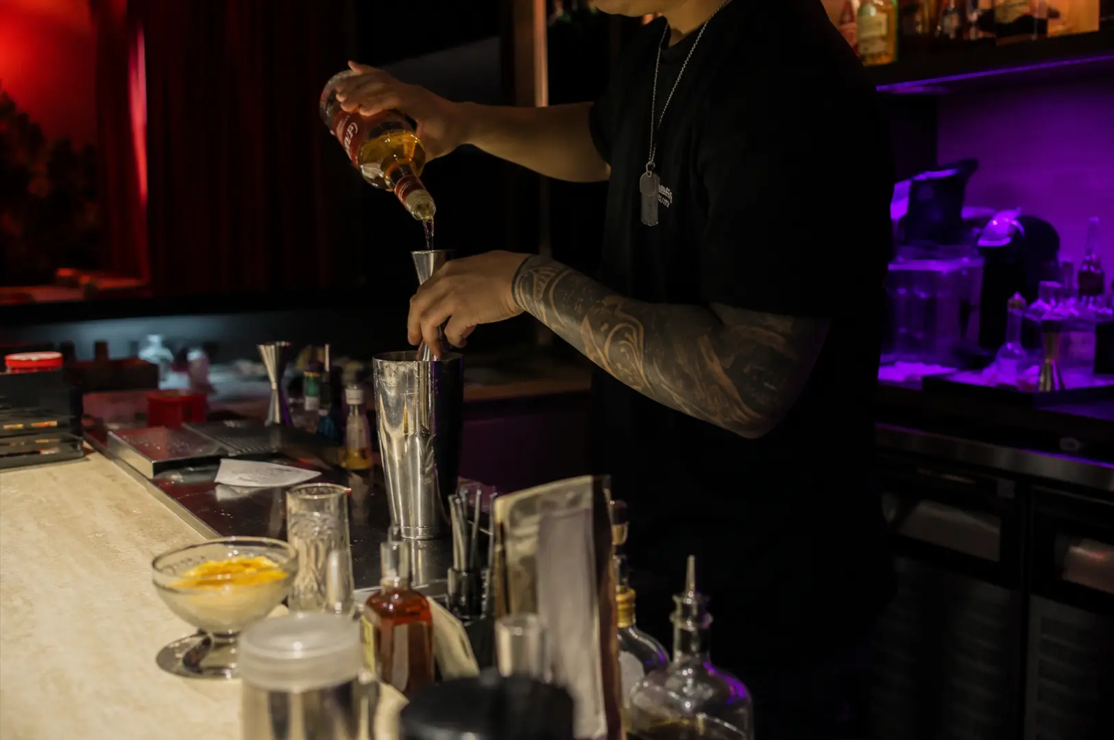Side view of an APT bartender carefully preparing a cocktail behind the bar, highlighting the attentive service and craft that define the café's evening experience.
