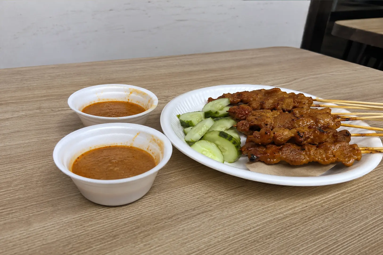 Grilled satay skewers with charred edges served with fresh cucumber slices and two small bowls of creamy peanut dipping sauce on a wooden table