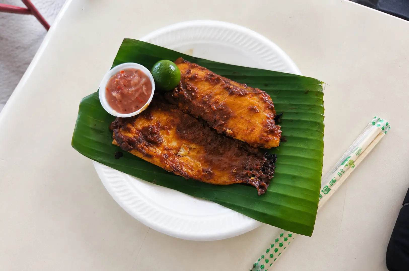 BBQ sambal stingray served on a banana leaf with a side of chili dip, calamansi lime, and chopsticks