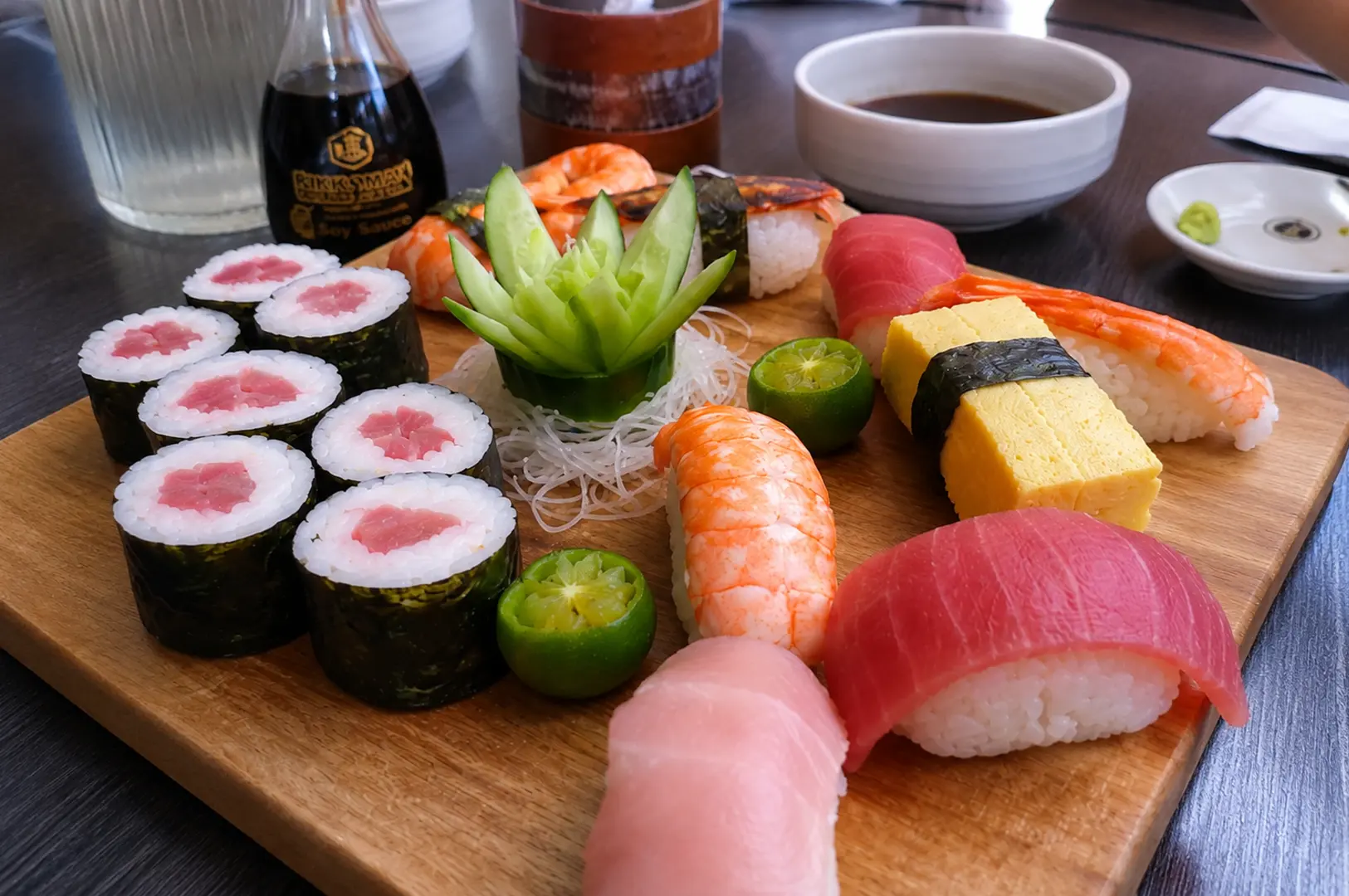 Assorted sushi platter with tuna rolls, nigiri, shrimp, tamago, and fresh garnishes on a wooden board.