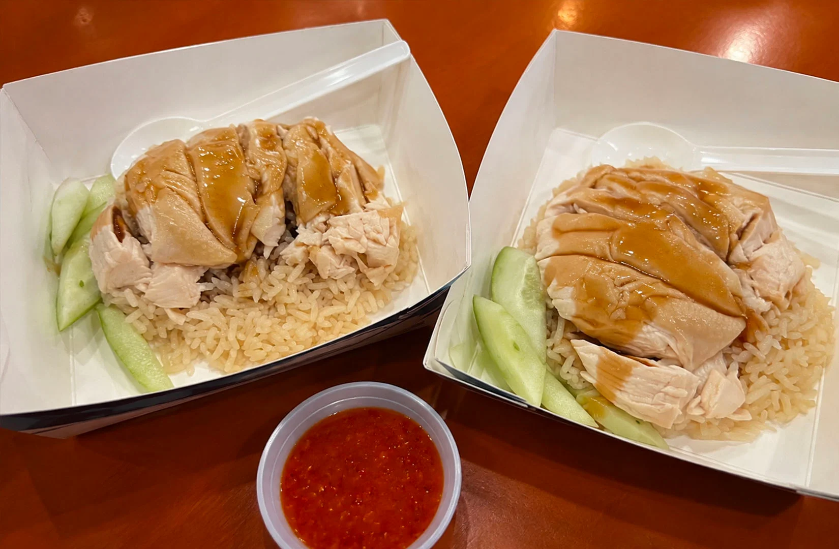 Two servings of Singapore Hainanese chicken rice topped with tender poached chicken, cucumber slices, soy sauce, and chili dip.
