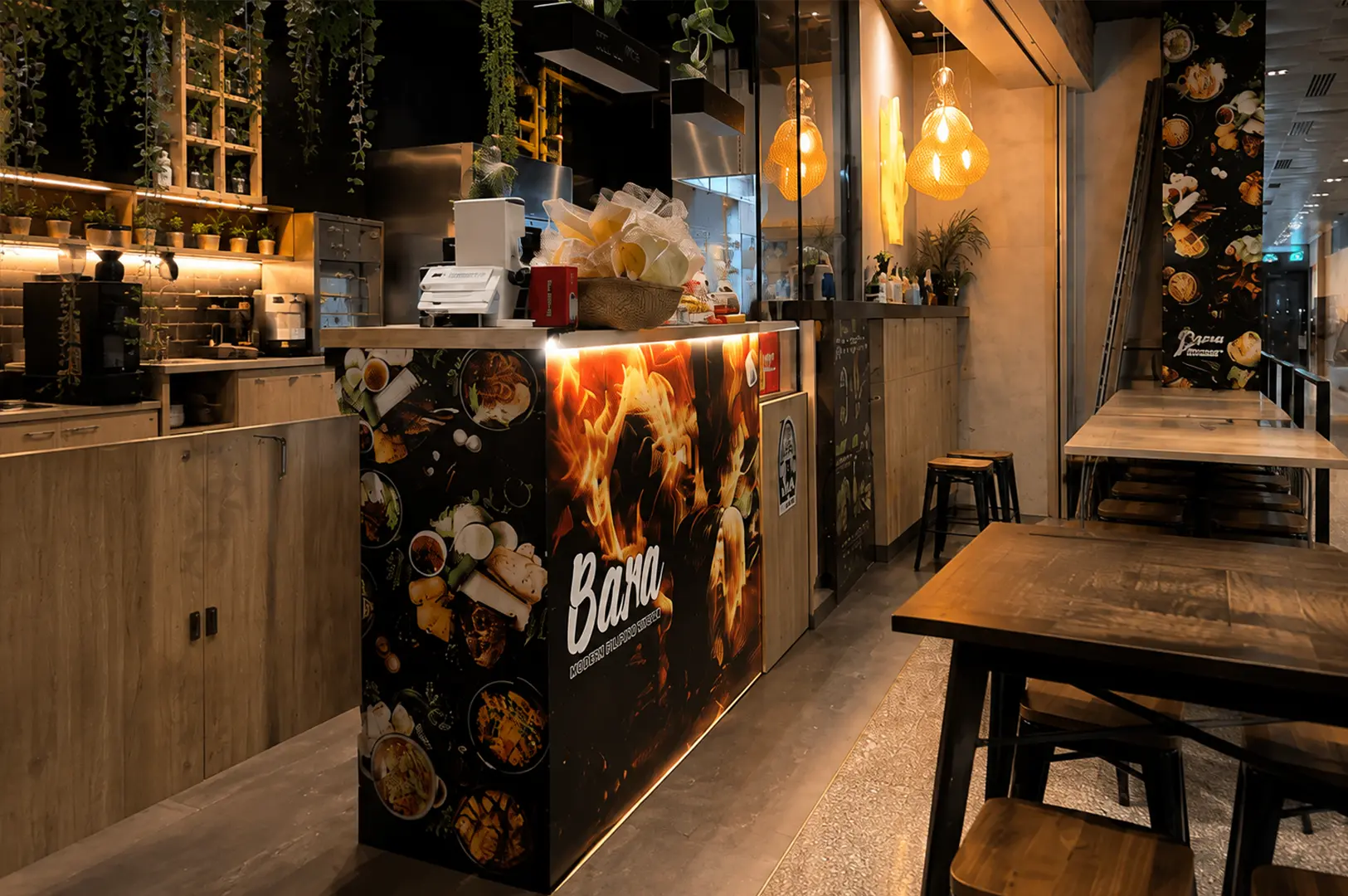 Warmly lit Bara Food Tanjong Pagar interior with wood furnishings, hanging plants, open kitchen counters, and dining tables.