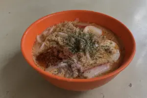 A bowl of creamy laksa on a simple table, topped with noodles, tofu, herbs, and chili paste.